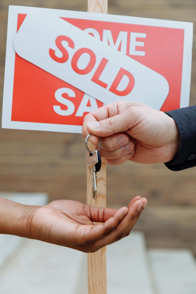 Hands exchanging house keys in front of a sold sign, symbolizing real estate success.