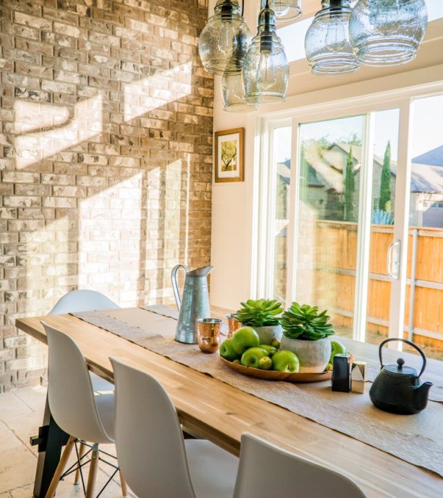 Sunlit dining room with contemporary decor and a rustic brick wall, featuring stylish lighting and a wooden table.